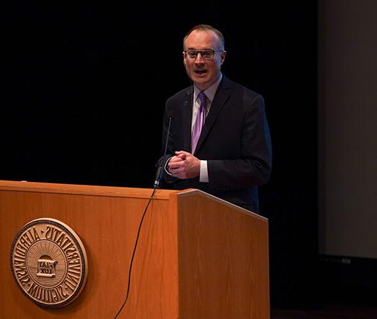 man with coat 和 tie, glasses, speaking at podium